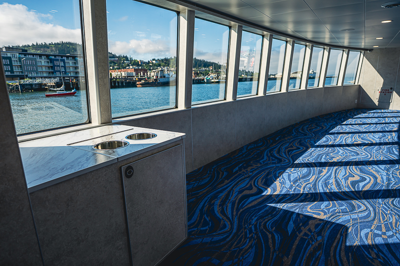 HBC_El Escudo_Marketing_Onboard_Web-10 Bright ferry interior with large windows, blue swirling carpet, and a marble counter with two cup holders, overlooking a harbor scene.