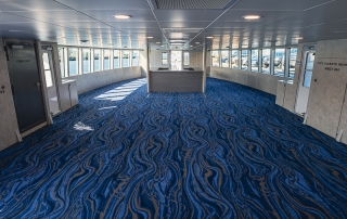 Interior of a ferry passenger lounge with blue wavy carpet, rows of windows, and a central service desk at the far end.