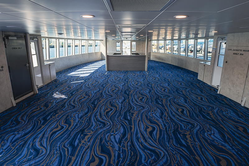 Interior of a ferry passenger lounge with blue wavy carpet, rows of windows, and a central service desk at the far end.