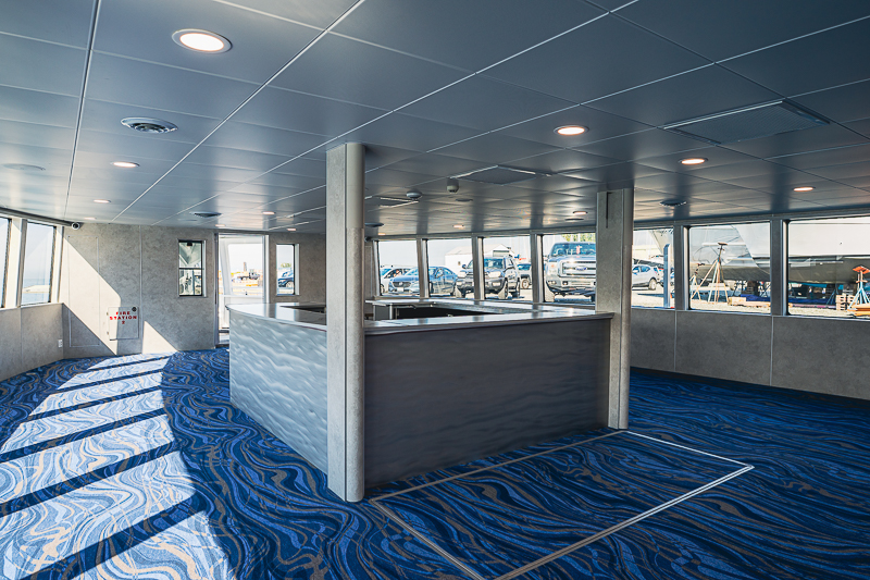 HBC_El Escudo_Marketing_Onboard_Web-12 Interior of a modern lounge with a curved blue patterned carpet, a central counter, and large windows showing vehicles outside.