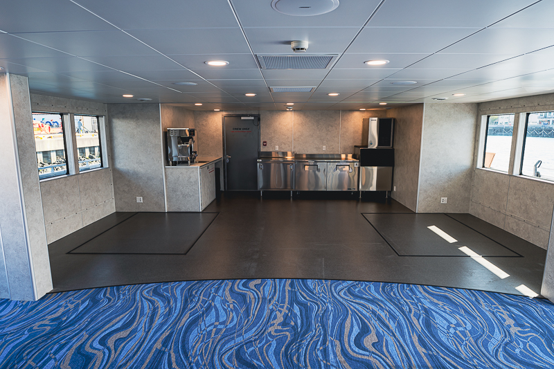HBC_El Escudo_Marketing_Onboard_Web-14 Interior of a ship's galley: stainless steel countertops and appliances along the back wall, coffee machine, and a clean, open prep area with windows on either side.