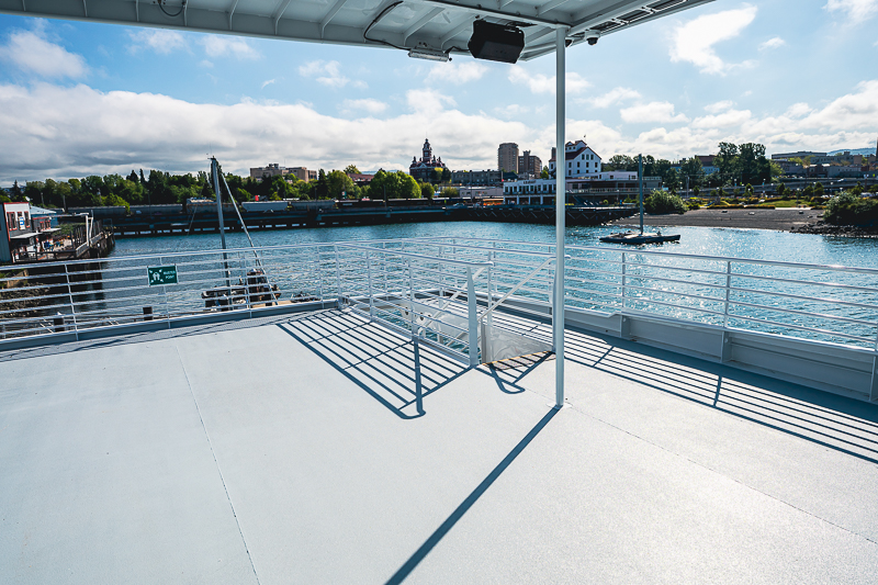 HBC_El Escudo_Marketing_Onboard_Web-15 View from a ship deck toward a river and city skyline, with railings and shadows on a bright day