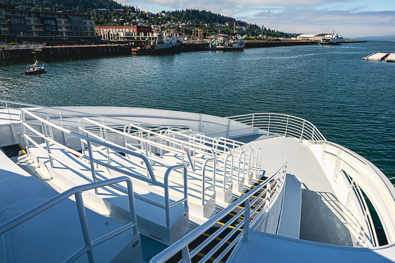 HBC_El Escudo_Marketing_Onboard_Web-16 White ship deck with safety rails and stairs, overlooking a harbor with boats and a coastal town in the background