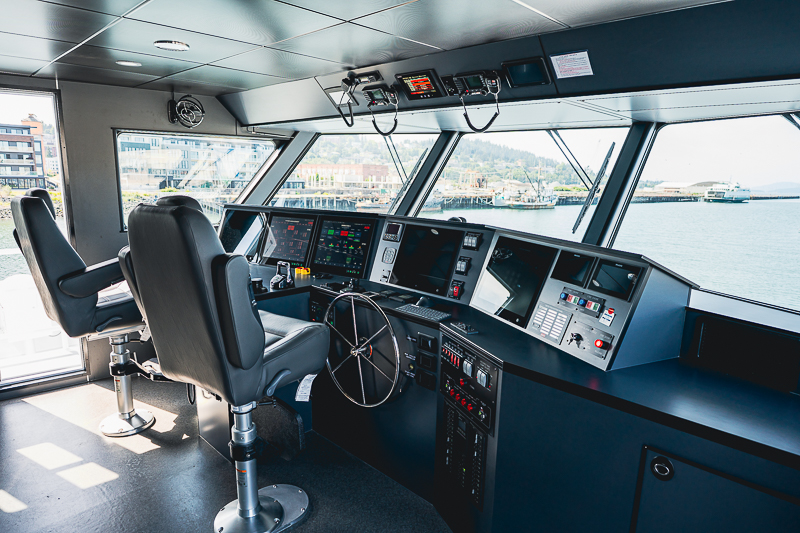 HBC_El Escudo_Marketing_Onboard_Web-17 Bridge of a ship with captain’s chair, steering wheel, and control panels overlooking a harbor