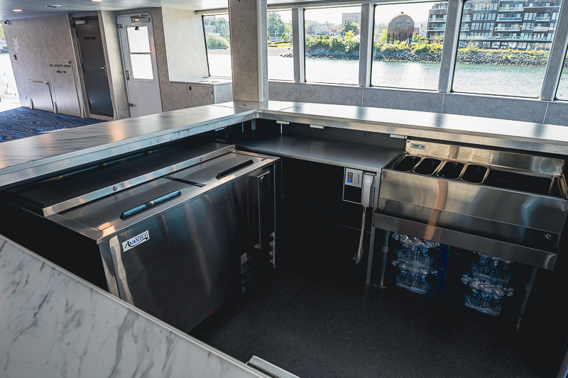 HBC_El Escudo_Marketing_Onboard_Web-2 Stainless steel beverage station with ice machines and a bottle cooler beneath a counter by large windows on a boat interior.