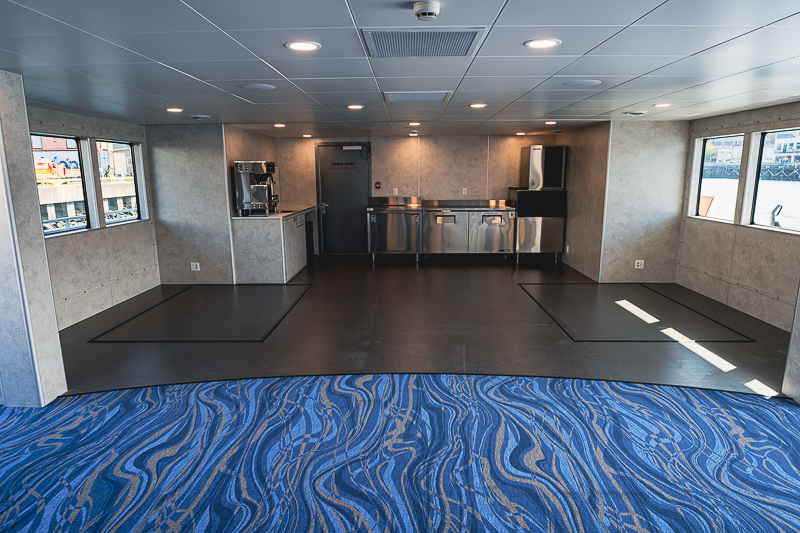 HBC_El Escudo_Marketing_Onboard_Web-4 Empty ship galley with stainless steel counters and appliances along the back wall, viewed from a foreground area with blue wavy carpet.
