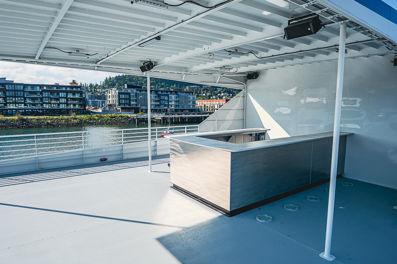 HBC_El Escudo_Marketing_Onboard_Web-7 Outdoor boat deck with a stainless bar counter under a covered area; railing along the water and modern buildings along a river in the background.
