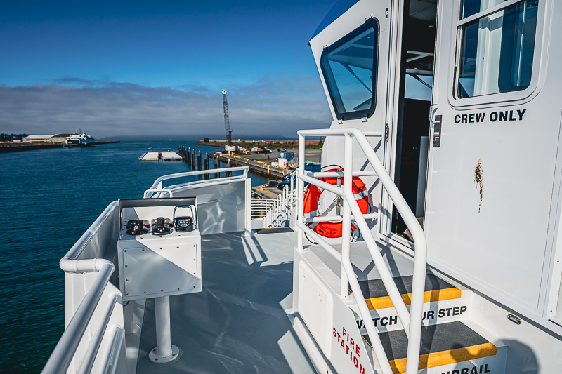 HBC_El Escudo_Marketing_Onboard_Web-8 Deck of a white vessel with a 'CREW ONLY' door, safety rails, and a life ring, overlooking a blue harbor with docks and calm water.
