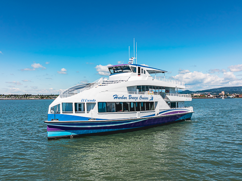 Large white and blue cruise boat named Harbor Breeze Cruises on calm harbor water with a distant shoreline.