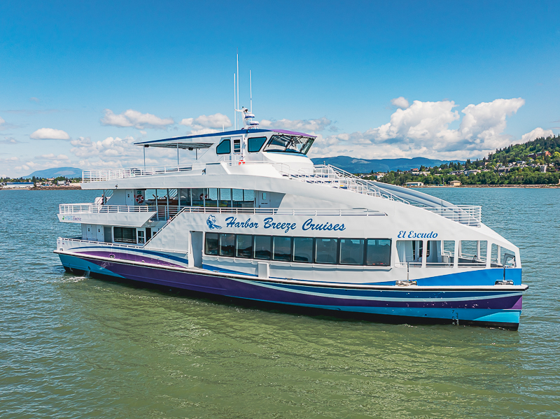 White and blue passenger cruise boat named Harbor Breeze Cruises on calm water with a shoreline and hills in the background.