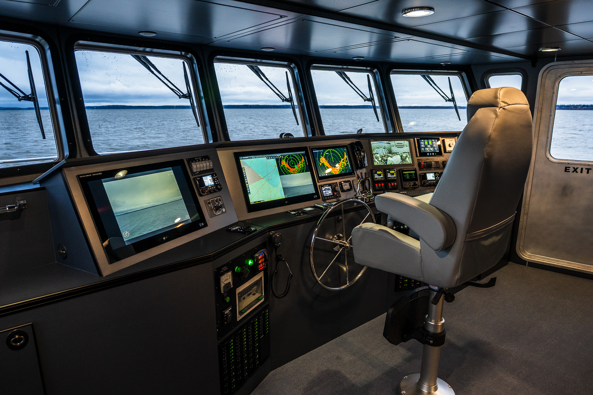 Bridge of a vessel with multiple navigation screens, a captain's chair, and water visible through windows.