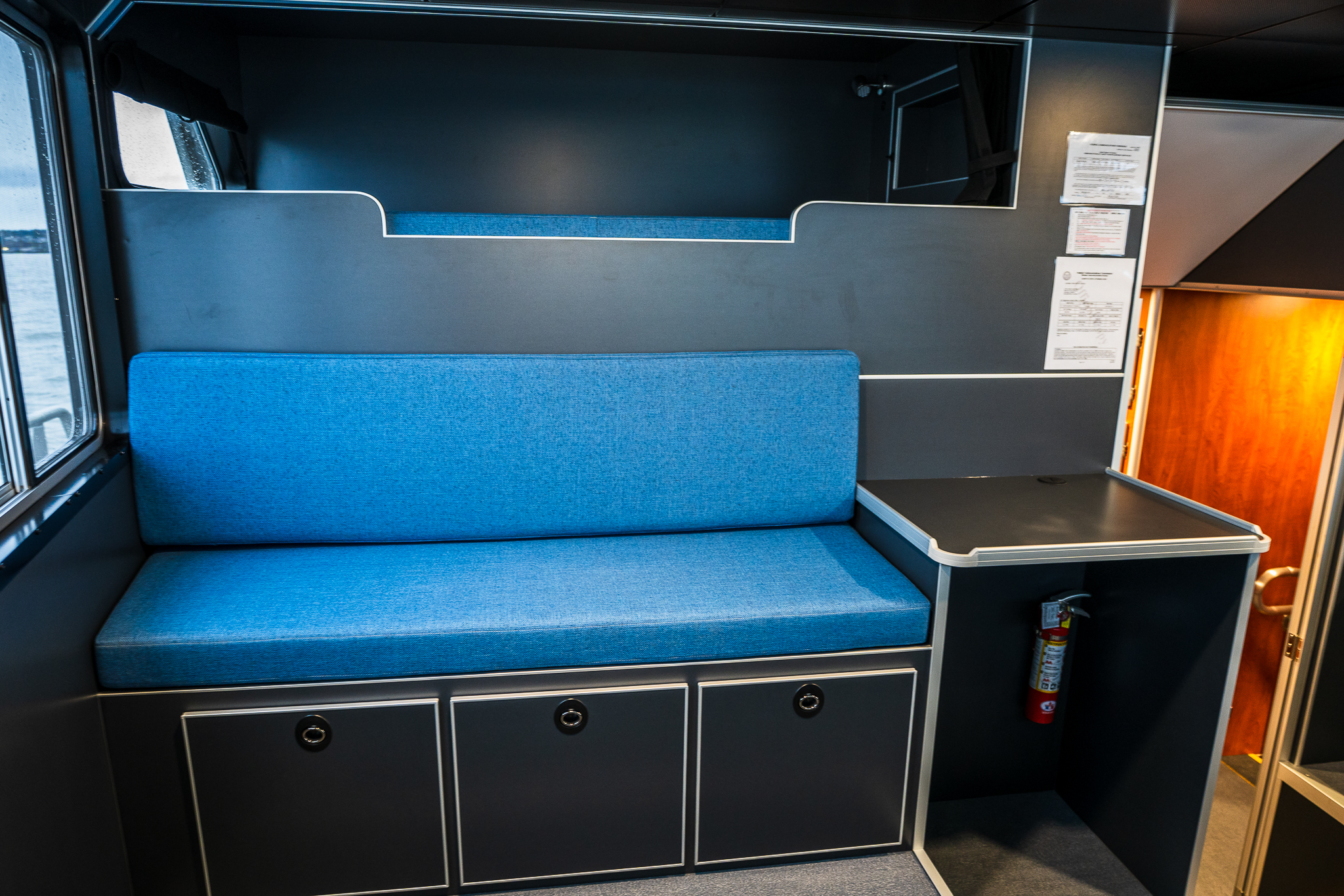 Blue upholstered bench seating and built-in storage lockers inside a train or bus cabin, with a fire extinguisher on the wall to the right.