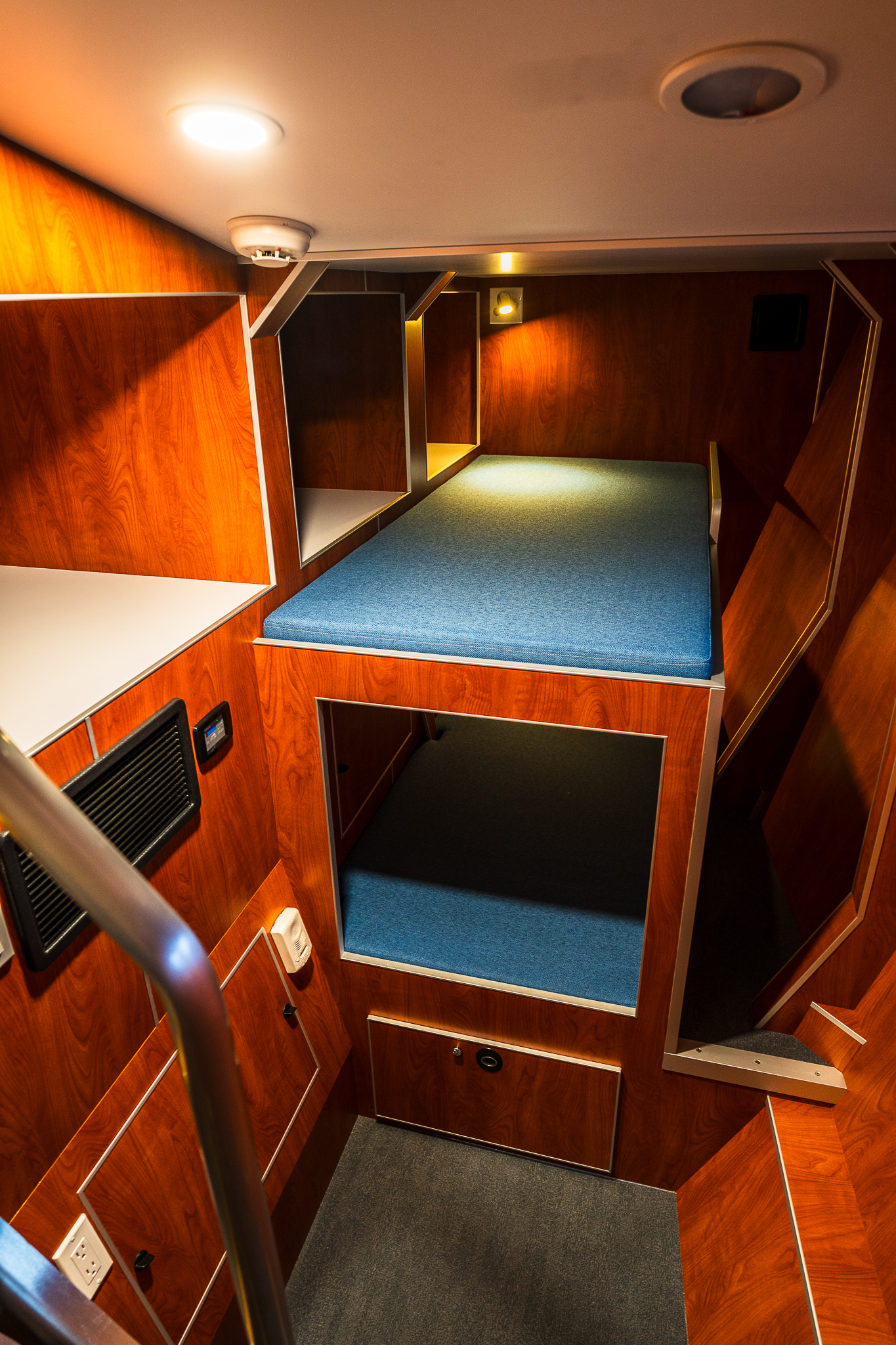 Wood-paneled sleeper cabin with a blue mattress on a raised bunk and small reading lights along the wall.