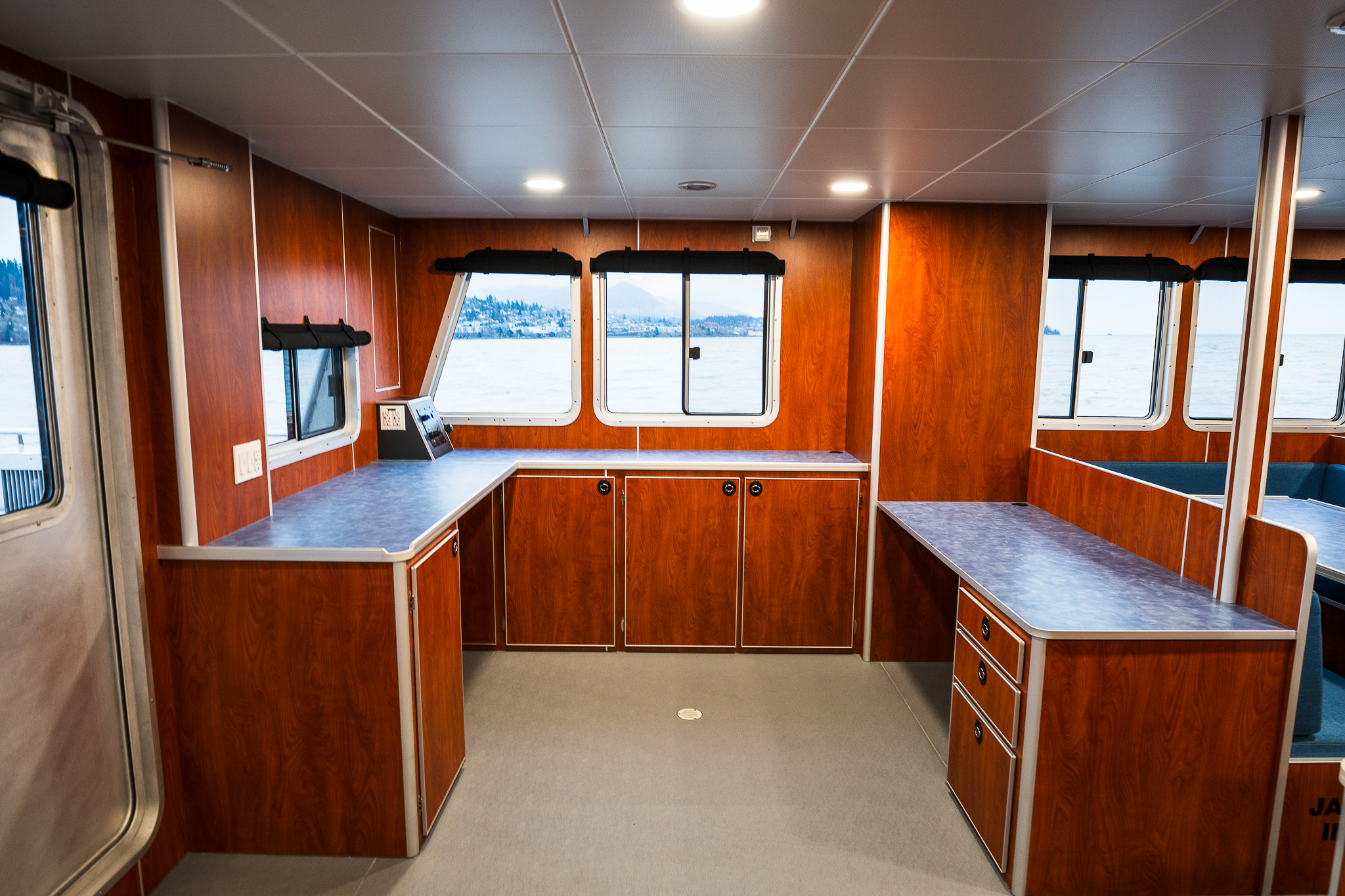 Wood-paneled boat galley with countertops, cabinets, and windows overlooking calm water outside