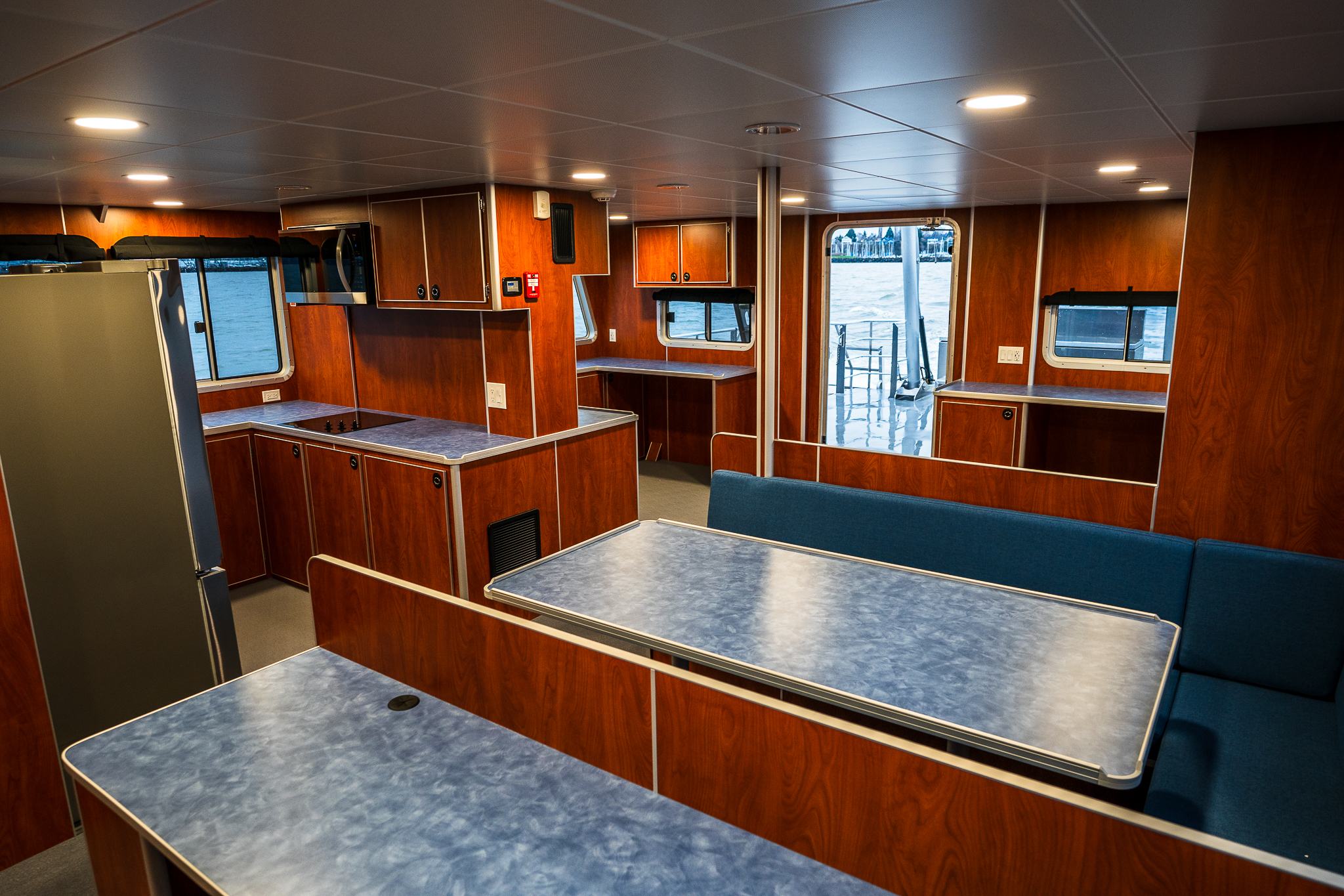Interior of a boat galley with dark wood cabinets, blue countertops, and windows showing water outside