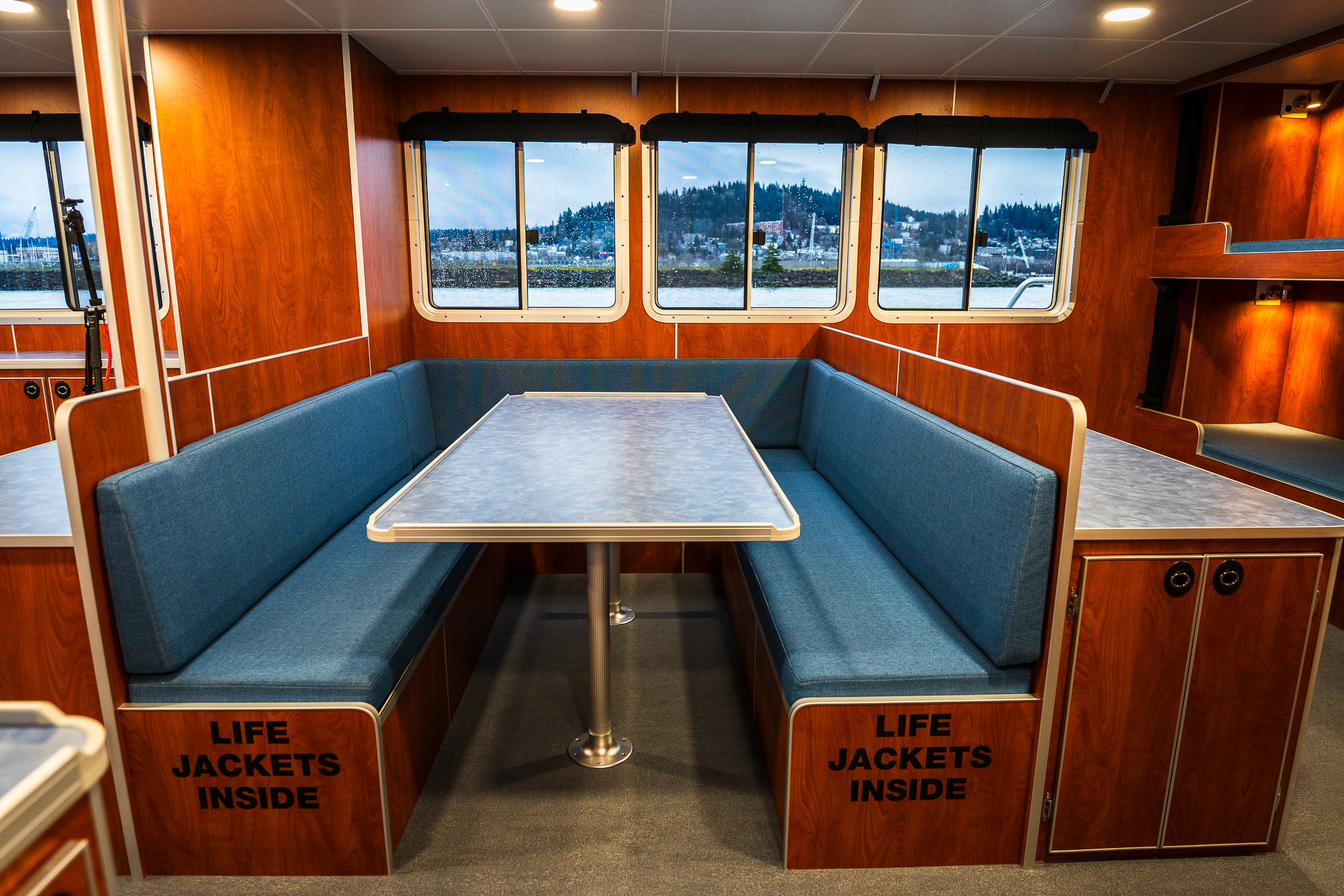 Interior of a wooden boat cabin with blue upholstered bench seating around a central metal table, and windows overlooking water and shoreline.