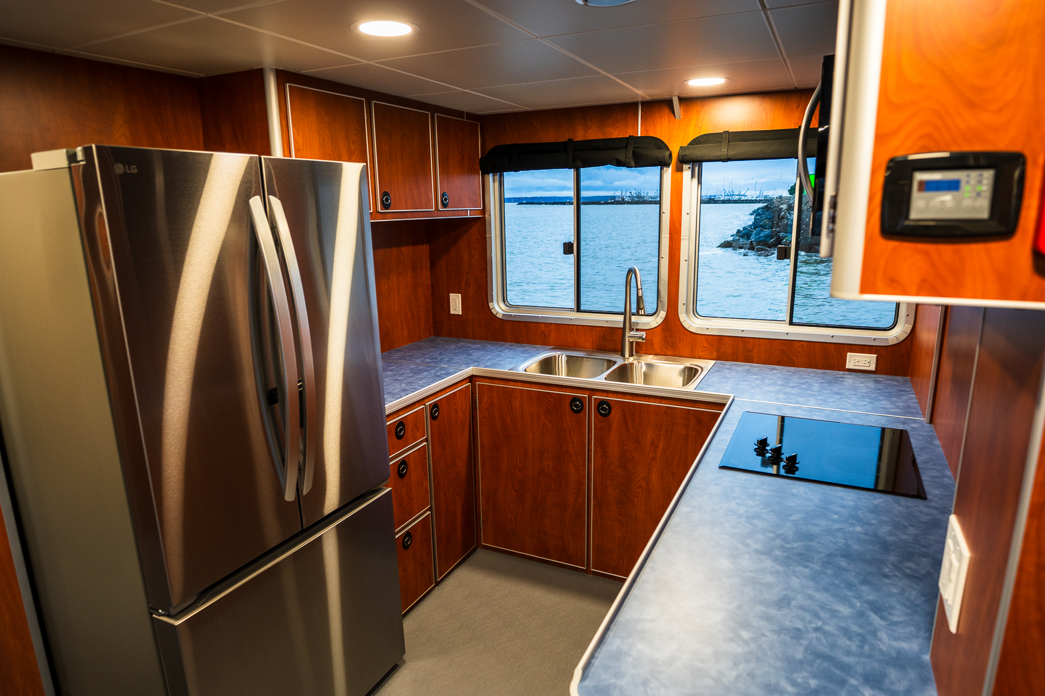 Wood-paneled galley on a boat with a stainless steel refrigerator, multiple cabinets, and a view of the water through the windows