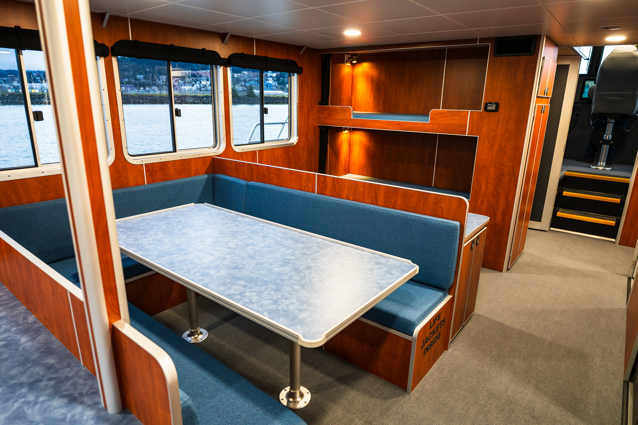 Interior of a boat cabin with blue cushioned bench seating around a light blue table and wood panel walls, windows looking out to water.
