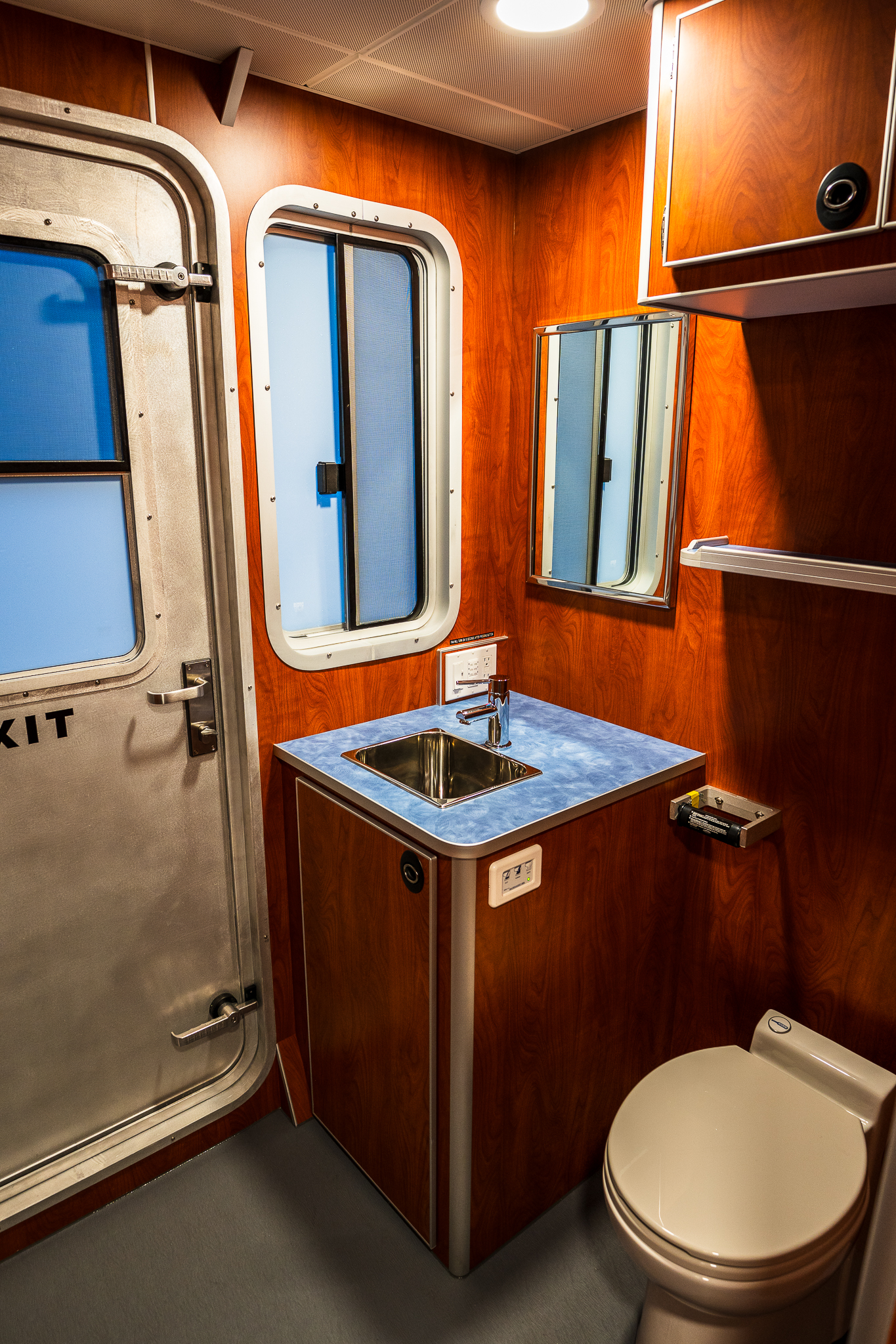 Compact train bathroom with wood paneling, stainless steel door, blue-tinted window, and blue countertop sink.