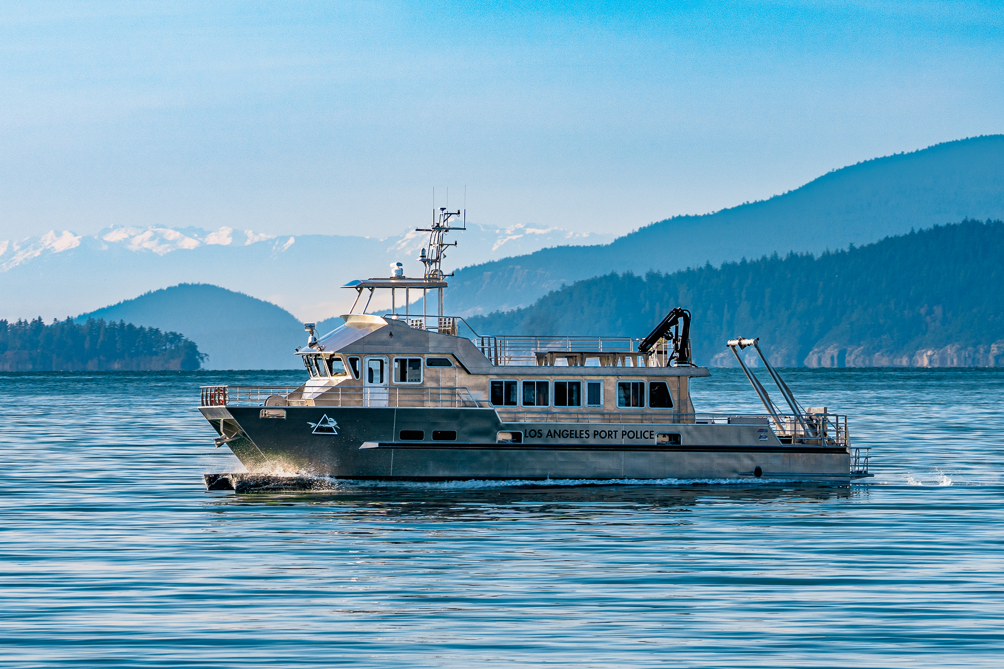 Port police patrol boat on calm blue water, with forested hills and snow-capped mountains in the distance, under a clear sky.