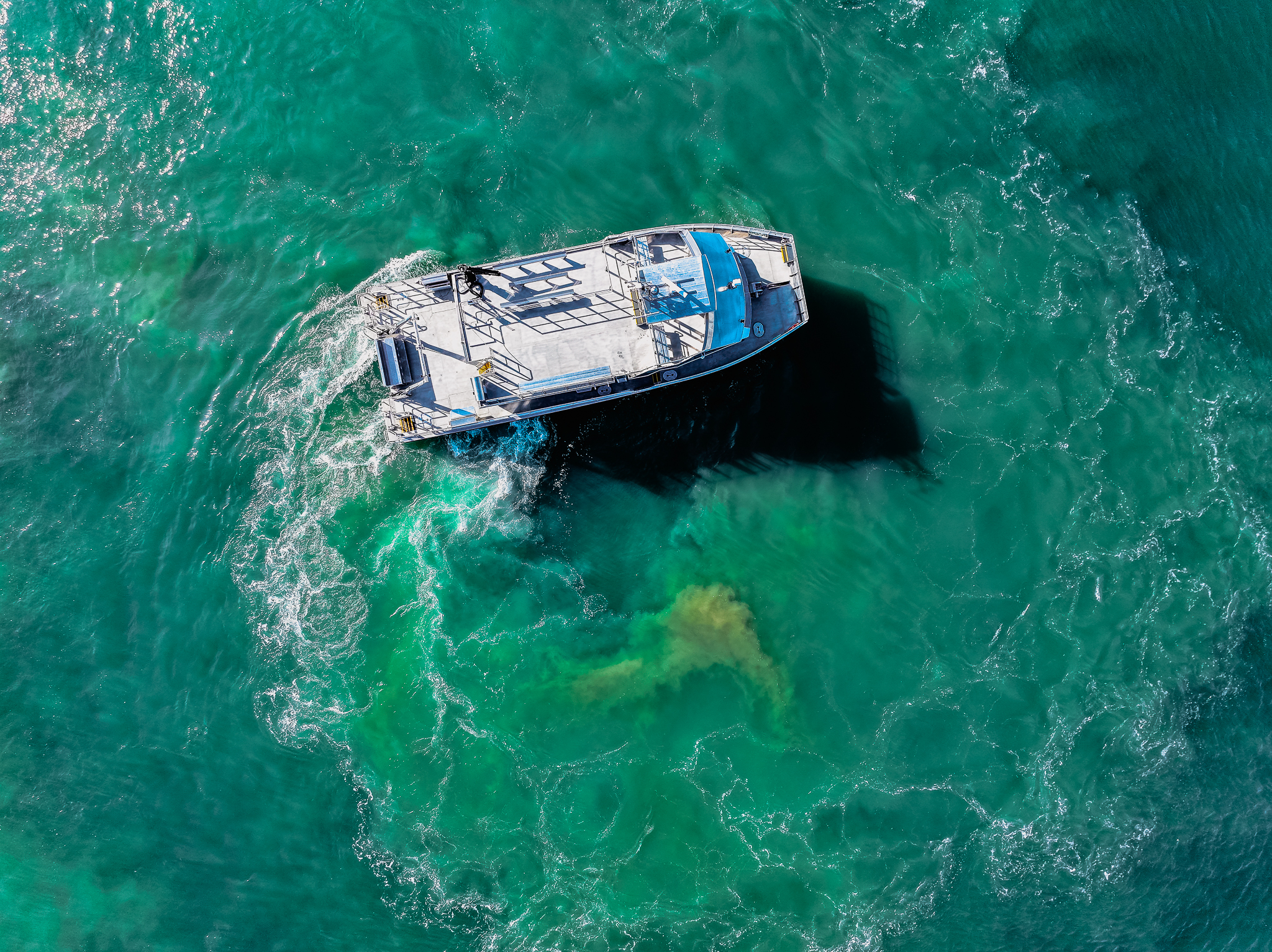 Aerial view of a white and blue boat sailing through turquoise water, with a dark shadow beneath it in the sea.