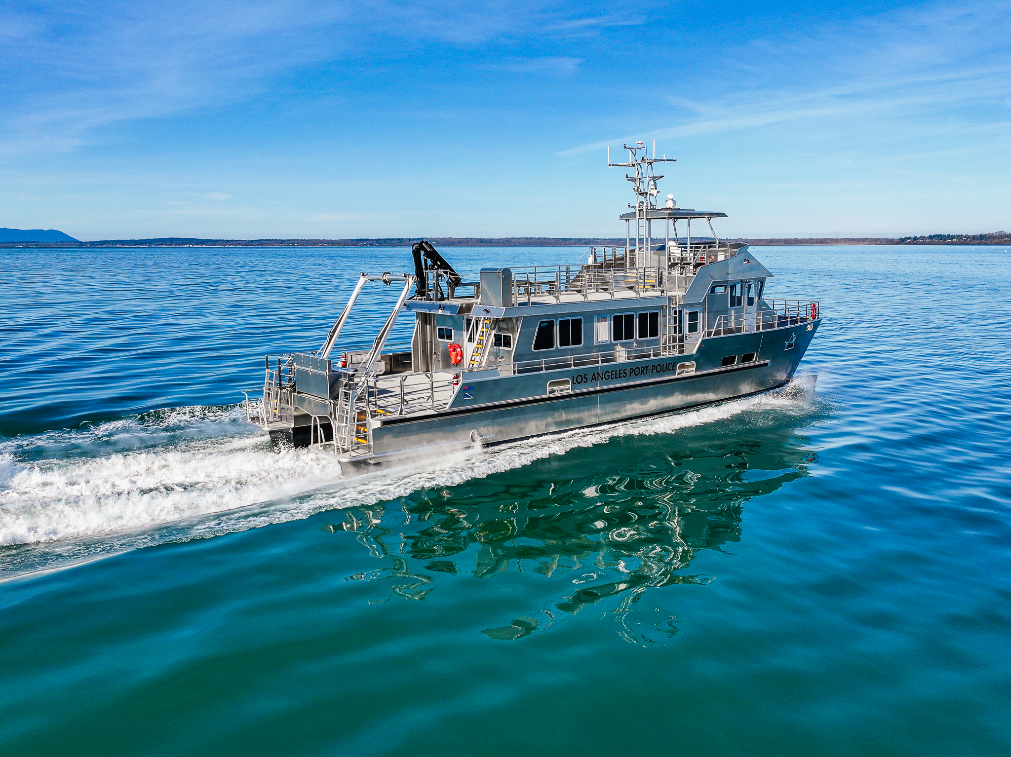 Port Police patrol boat named Los Angeles Port Police speeds across blue harbor water, leaving white wake with a distant shoreline on the horizon.